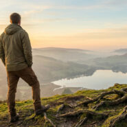 Homme de dos au sommet d'une colline, contemplant une vallée avec lac et montagnes brumeuses au coucher du soleil. Racines d'arbre apparentes.