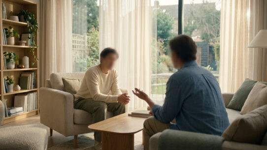 Deux personnes en conversation thérapeutique dans un salon lumineux avec bibliothèque et vue sur jardin. Ambiance sereine.