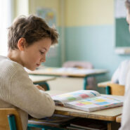Un jeune élève concentré sur un livre, avec une main adulte posée sur son épaule, dans une salle de classe lumineuse.