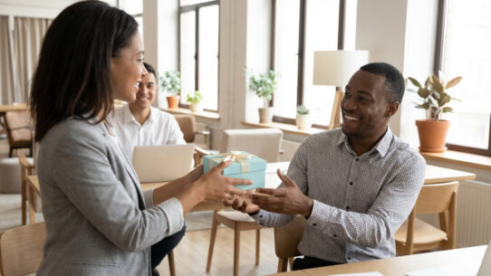 Une femme offre un cadeau à un collègue souriant dans un bureau moderne. Le geste symbolise le bien-être et la reconnaissance.
