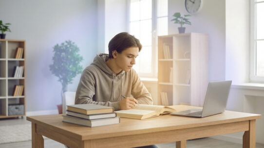 Un jeune homme étudie à son bureau, entouré de livres et d'un ordinateur portable, sous une lumière naturelle éclatante.