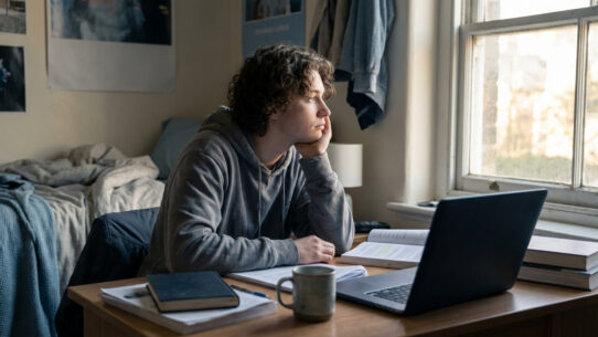 Jeune homme pensif et triste, cheveux bouclés, dans sa chambre d'étudiant désordonnée, fixant la fenêtre. Livres et laptop sur le bureau.