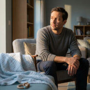 Homme pensif assis dans un salon, regardant par la fenêtre. Un plaid de bébé et un jouet sont sur le canapé à côté de lui.