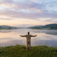 Personne les bras ouverts face à un lac brumeux au lever du soleil, des orbes lumineux flottent sur l'eau.