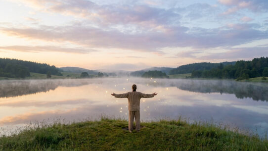 Personne les bras ouverts face à un lac brumeux au lever du soleil, des orbes lumineux flottent sur l'eau.