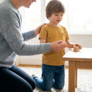 Une mère s'agenouille doucement près de son jeune fils pour le guider dans une activité calme, dans un intérieur lumineux.