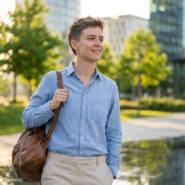 Jeune personne souriante marchant dans un parc urbain, portant une chemise bleue et un sac en cuir, sous une lumière douce.