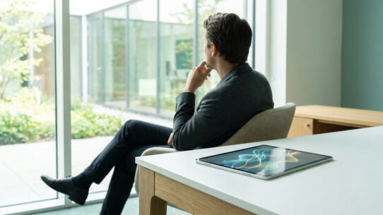 Un homme en costume assis dans un cabinet lumineux regarde par la fenêtre, une tablette avec des ondes visuelles à côté.