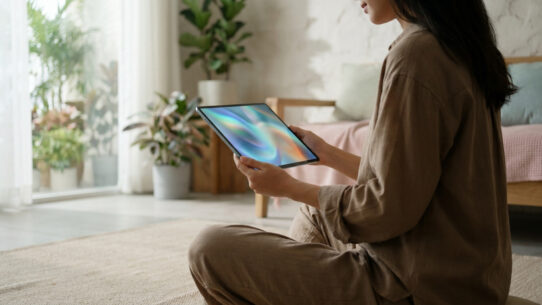 Une femme assise en tailleur chez elle tenant une tablette affichant un fond d'écran abstrait aux couleurs apaisantes.