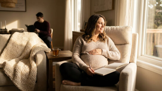 Une femme enceinte assise dans un fauteuil près d'une fenêtre, tenant un carnet, avec son partenaire en arrière-plan.