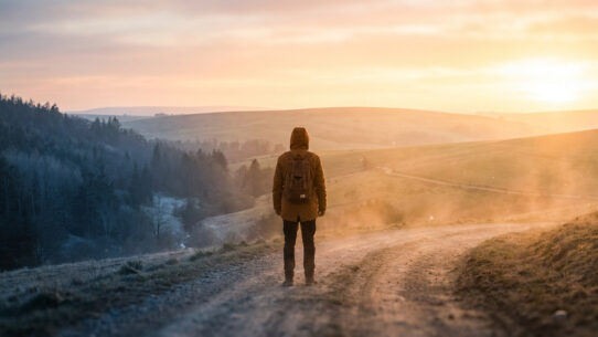 Une personne avec un sac à dos marchant sur un chemin de terre vers un paysage ensoleillé.