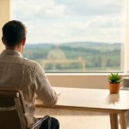 Un homme assis à son bureau regarde par la fenêtre un paysage paisible, évoquant une pause pour éviter le burn-out.